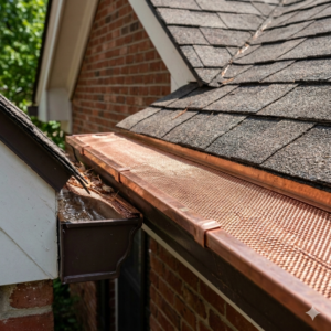 A close-up product image showing a copper mesh gutter guard in a real-world application. The bright, new copper wire mesh is seamlessly fitted onto the gutter system, effectively blocking debris and autumn leaves. The background features dark grey roof shingles and a classic red brick wall.