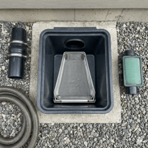 A real-world application setup of a Western-style residential rainwater harvesting and filtration system on a gravel ground. In the center, a trapezoidal stainless steel rainwater filter basket with its metal bracket is installed inside a rectangular black plastic catch basin. To the left of the basin, a black drainage coupling pipe and a coiled grey corrugated hose are placed neatly; to the right, there is a pipe inline filter with a green frame. Rainwater Filter Basket Bracket