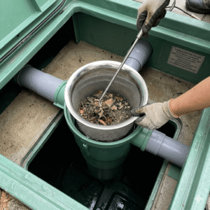 A real-world photo showcasing a European/American style residential underground pre-installed rainwater harvesting and filtration system. The green plastic storage tank's inspection hatch is open, revealing the internal pipe connections. A technician wearing protective gloves uses a metal hook handle to portably lift or inspect the stainless steel perforated mesh basket filter, which contains collected fallen leaves inside. Rainwater Filter Insert