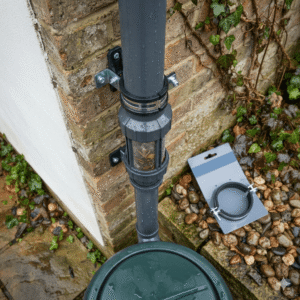 A close-up, top-down view of a grey downpipe with a rainwater harvesting filter installed on a brick wall. A secure metal Rainwater Pipe Clamp fixes the pipe to the wall, while a separate, unused Rainwater Pipe Clamp with rubber lining sits on its packaging nearby on the ground.